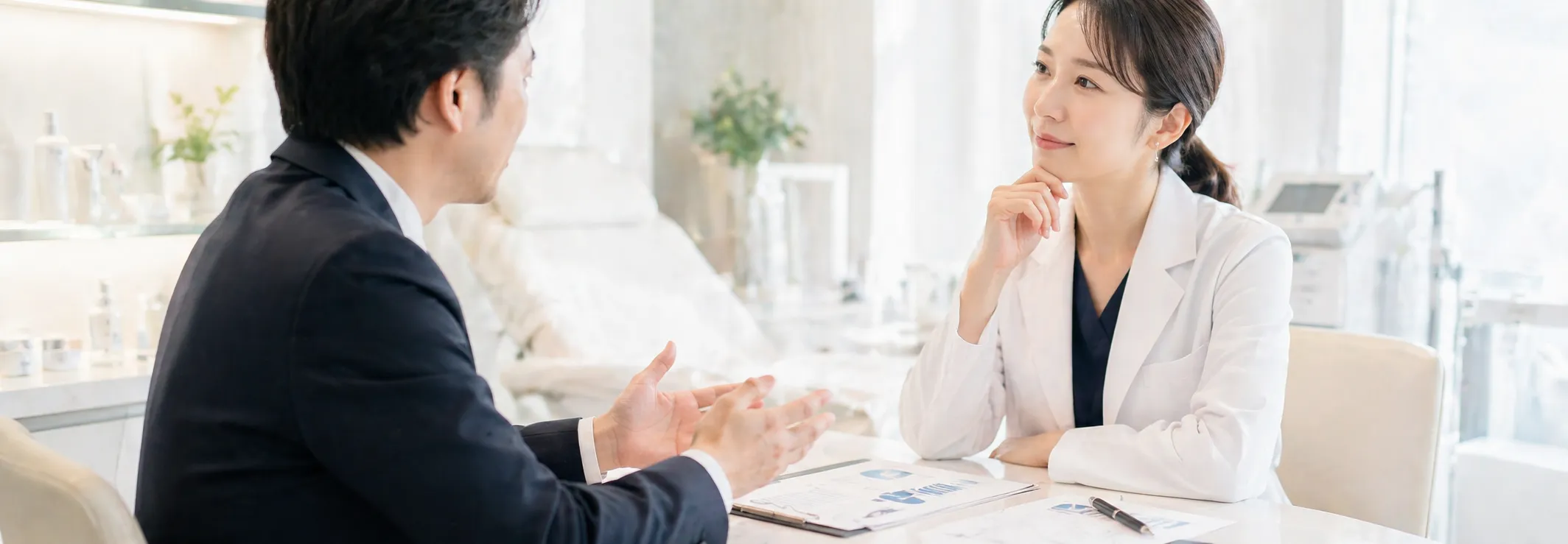 明るいオフィスで、男性と女性のビジネスパーソンが会議テーブルに向かい合い、書類とペンを前に置いて打ち合わせをしている。背景には観葉植物と大きな窓がある